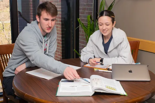 Two students review a textbook together while studying at a table with their laptops in the library