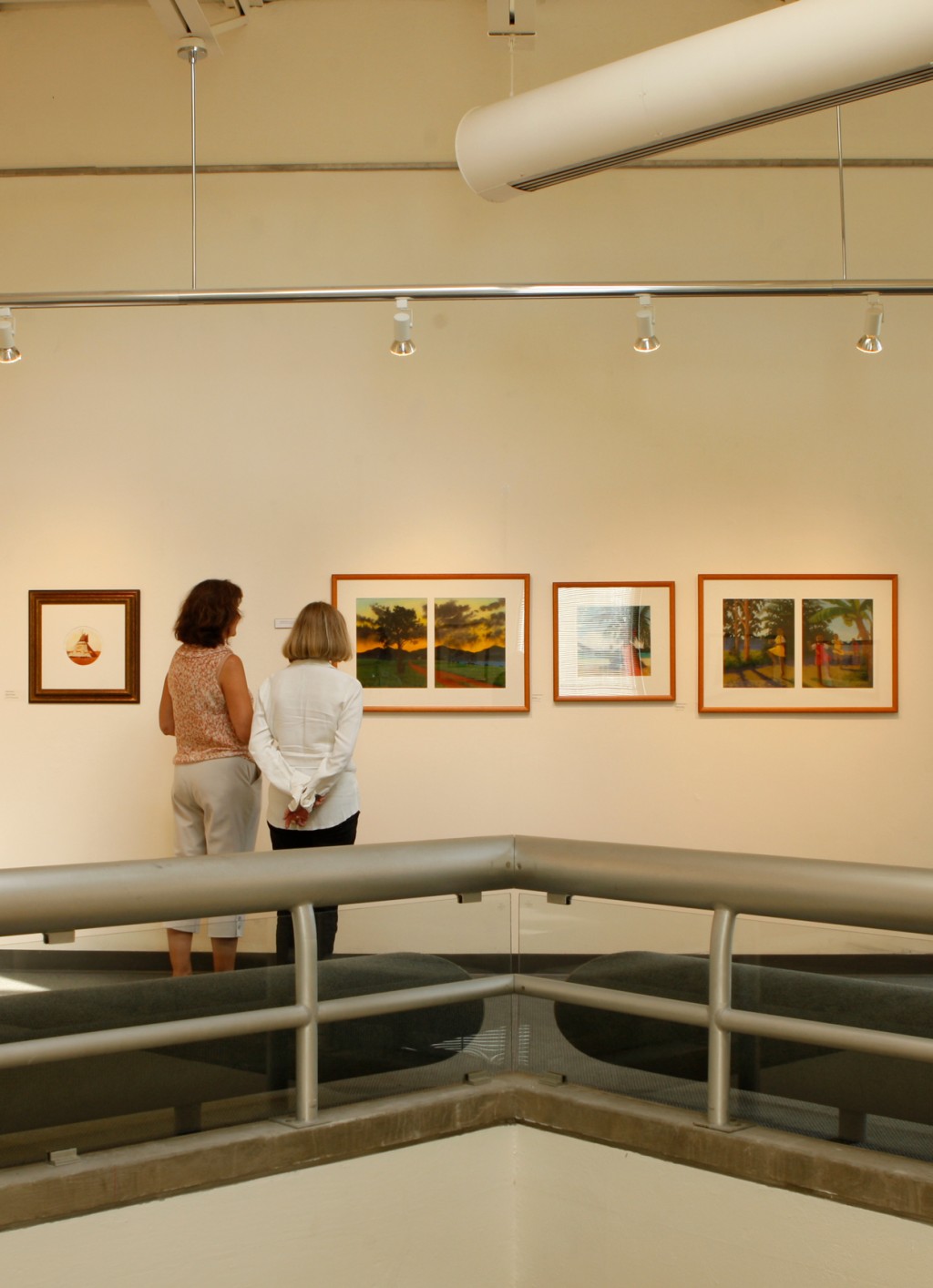 Two women look at a row of framed paintings hanging on a white wall in the Portland Campus art gallery