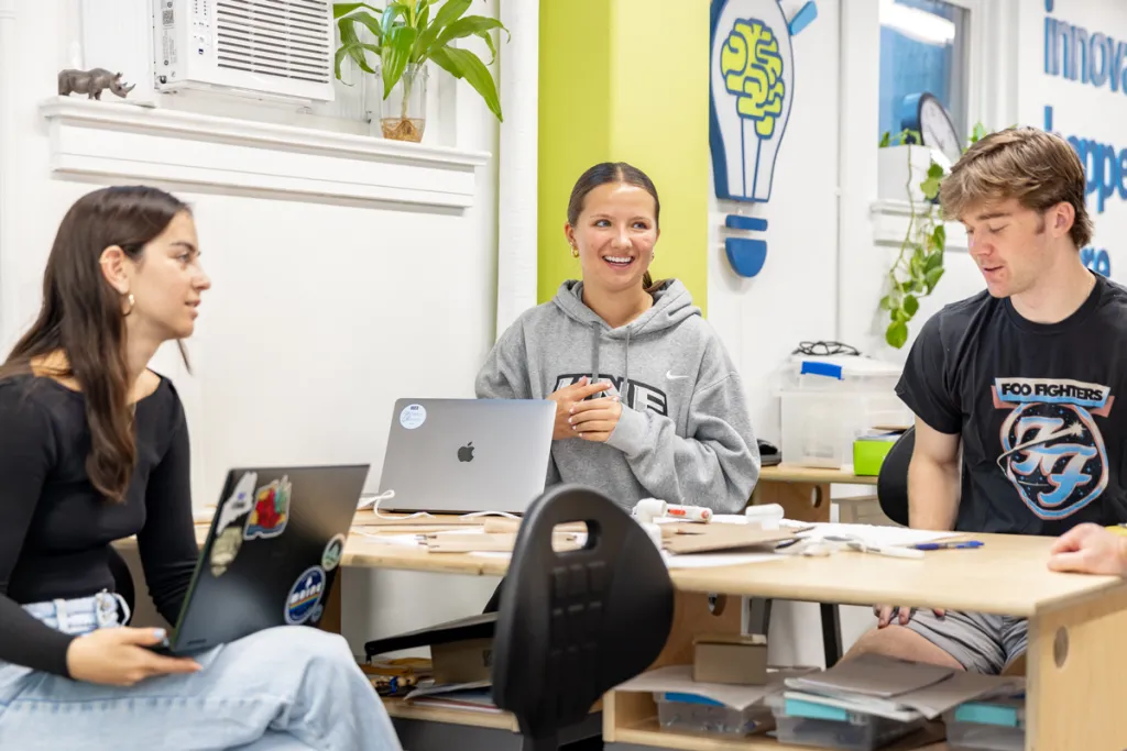 Three students talk to each other while sitting at a shared desk with their laptops