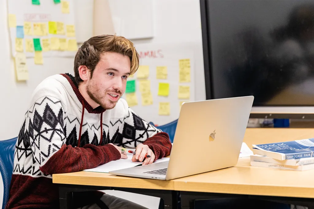 A student sits at their laptop in front of a whiteboard with several colorful post-its