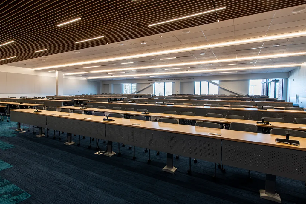A modern lecture hall with long, light-wooden tables, blue chairs, blue printed carpet, and wooden slat ceiling