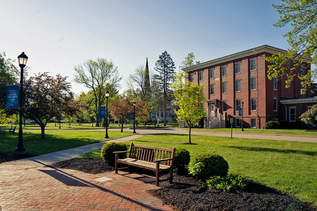 A wooden bench sits on a brick path in front of a three-story red brick building on a sunny day, surrounded by green trees and grass