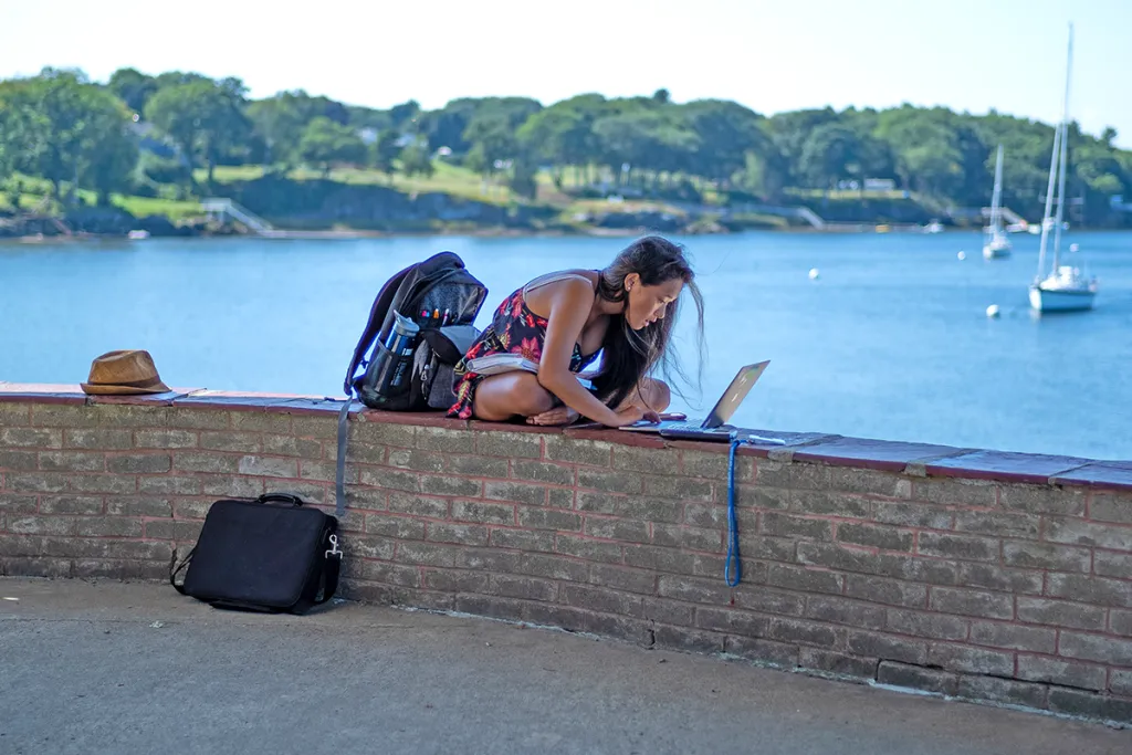 A student studies from their laptop while sitting on a brick wall in front of an ocean, filled with boats and surrounded by green trees