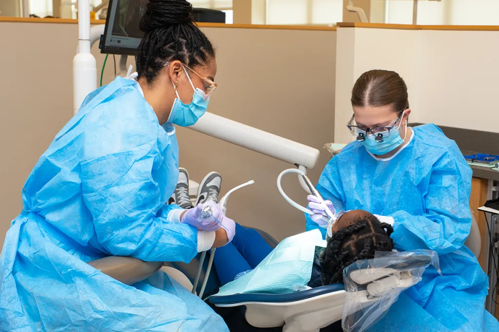 Two College of Dental Medicine students in blue scrubs check a child's patient teeth in the Oral Health Center