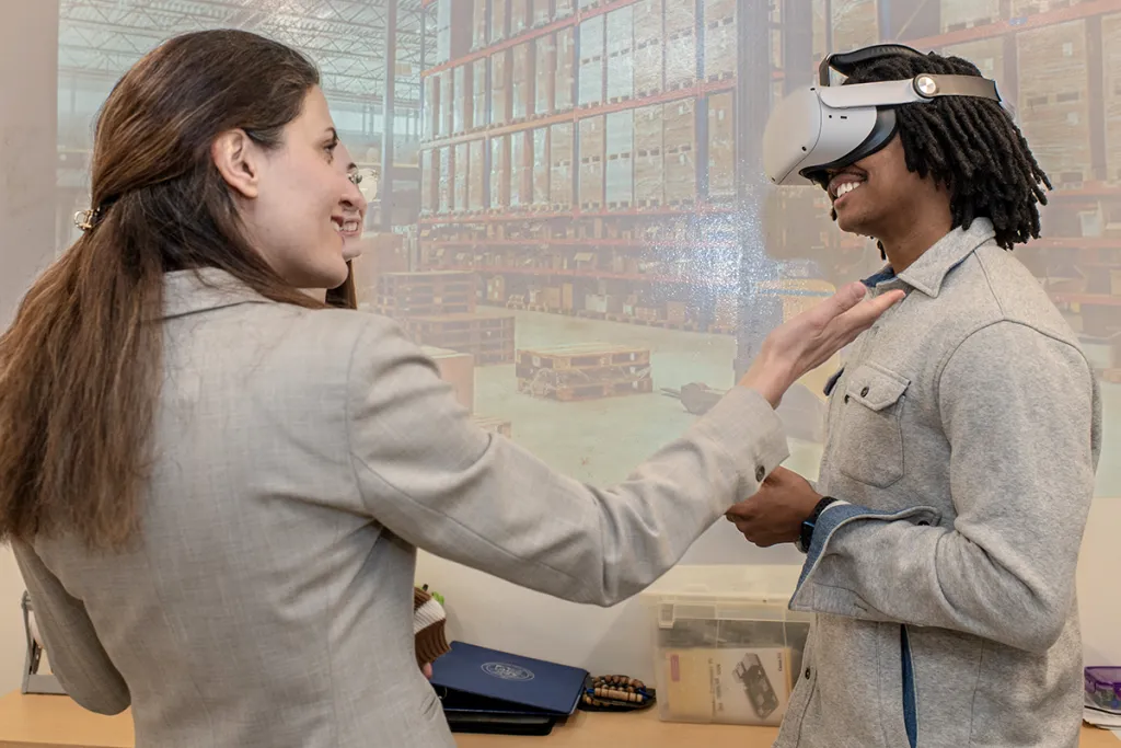 A professor and a student wearing a V R headset talk to each other in front of a projected image of a supply warehouse