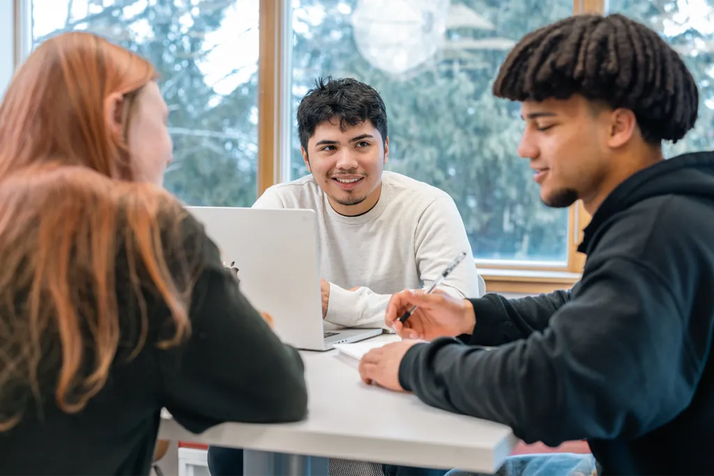 Three students sit around a small table and study together with textbooks and their laptop