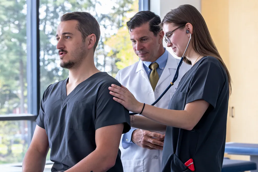 A physician assistant professor assists a student as they use a stethoscope to listen to the cardiac sounds of another student