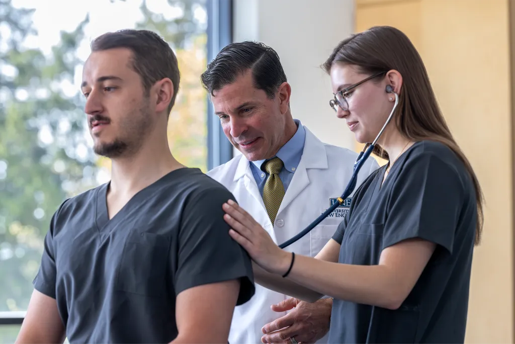 A physician assistant student practices listening to lung sounds with a stethoscope on another student as their professor mentors