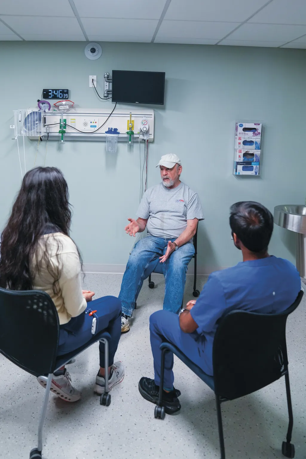 Michael, a patient actor wearing a ball cap, speaks to an interprofessional student team including Jasmin Gil, Ben Jones, Andrii Sokolov, and Varun Kota seated in a circle in a simulation room.