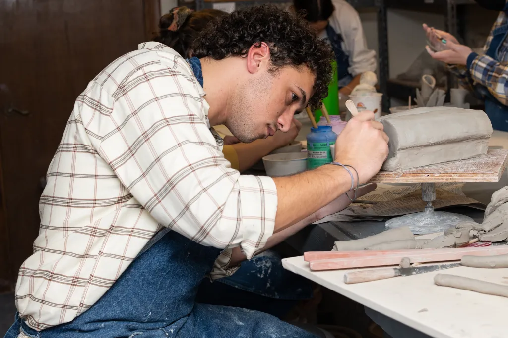 Art student focused on hand-building a ceramic piece at a studio workbench surrounded by clay tools and materials.