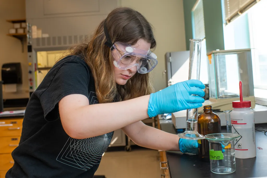Chemistry student wearing safety goggles and blue gloves carefully measuring liquid with a dropper into a beaker during a laboratory experiment.