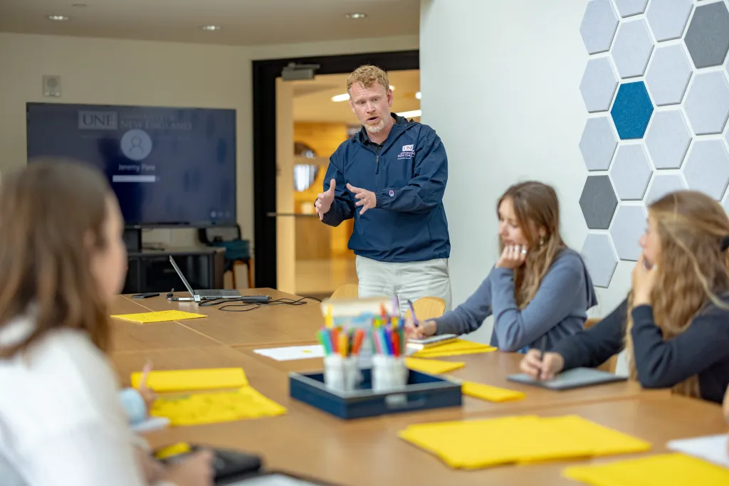 Professor Pare leads a discussion with students seated around a conference table with yellow notepads, a presentation screen visible in the background.