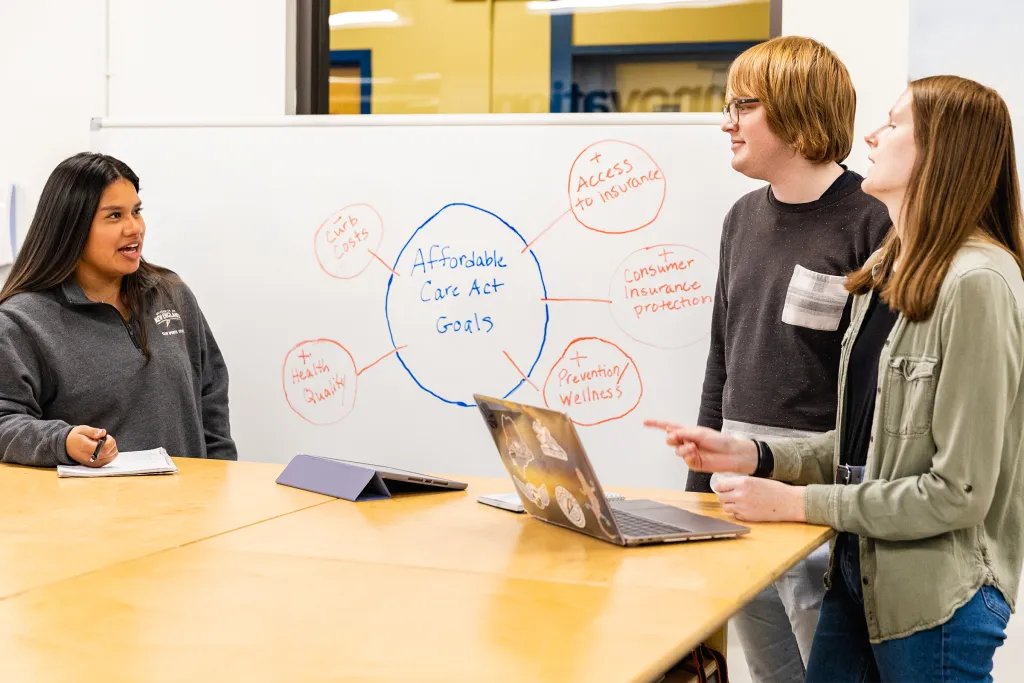 Three students presenting their Affordable Care Act research at a whiteboard showing a concept map with goals including health equity, access to insurance, and consumer protection.