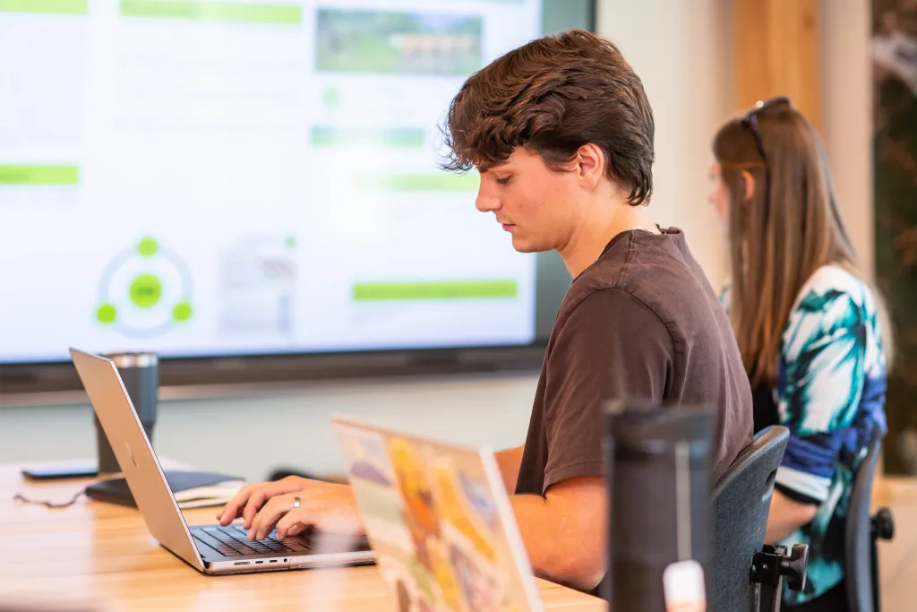 Students collaborating at laptops in a modern study space with a large presentation screen visible in the background.