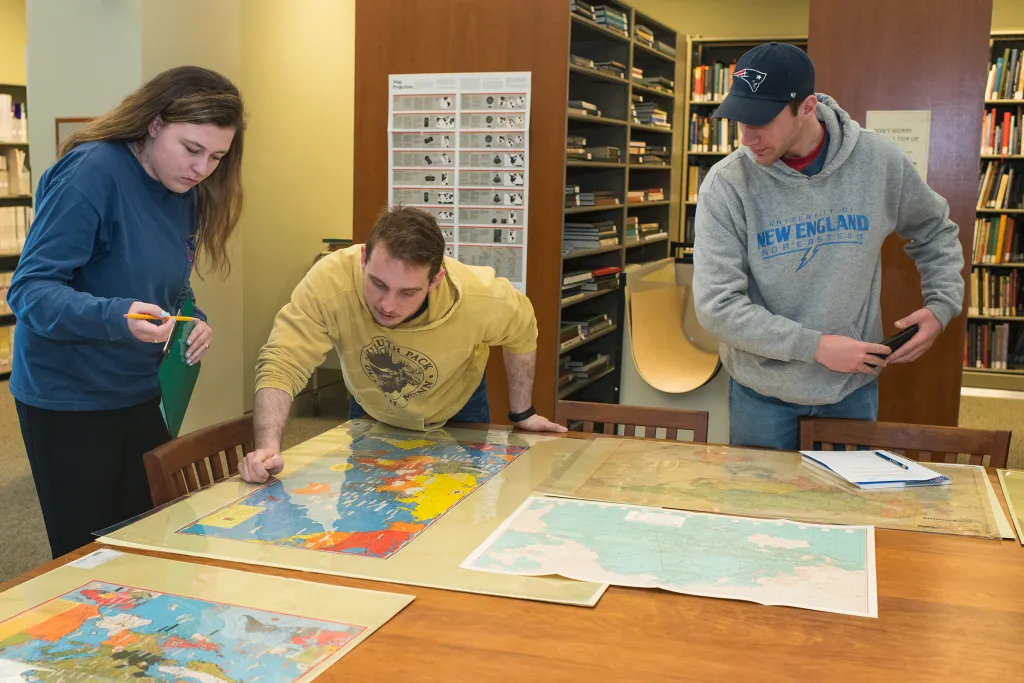 Three students examining historical maps and documents on a large table in a library archive room with map storage visible in the background.