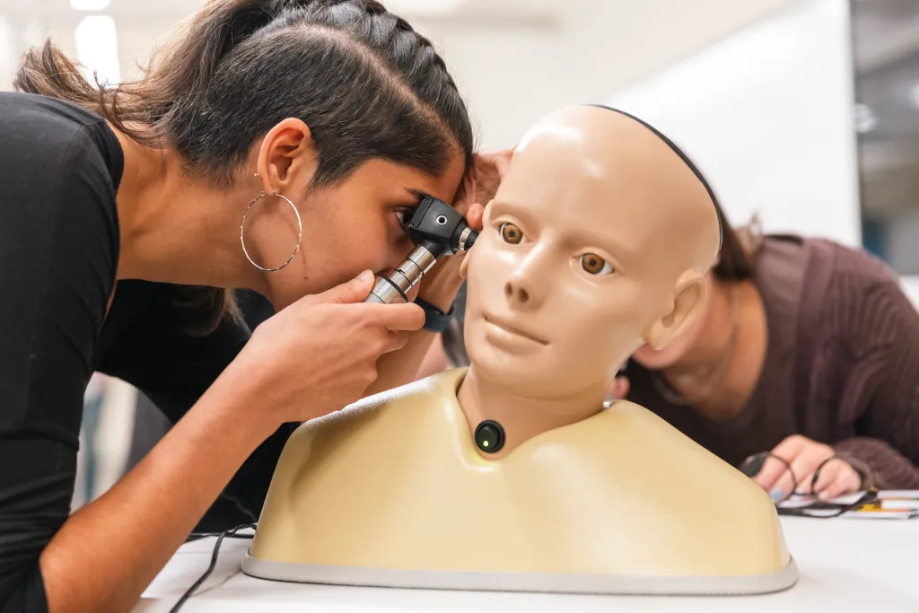 U N E nursing student using an otoscope to examine a simulated patient's ear during a clinical skills practice session.