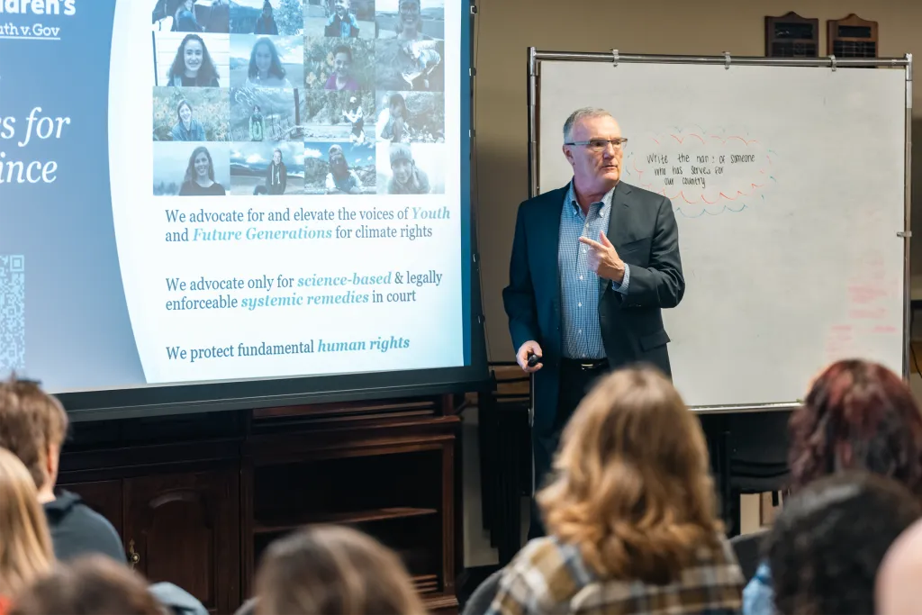 A speaker presenting to political science students about climate rights advocacy for youth and future generations, with a projection screen showing organizational mission statements.