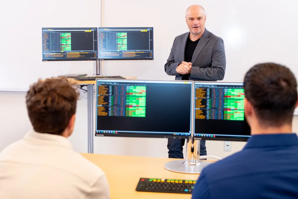 College of Business Dean Norm O’Reilly speaks to two students in a trading lab with multiple monitors displaying market data.
