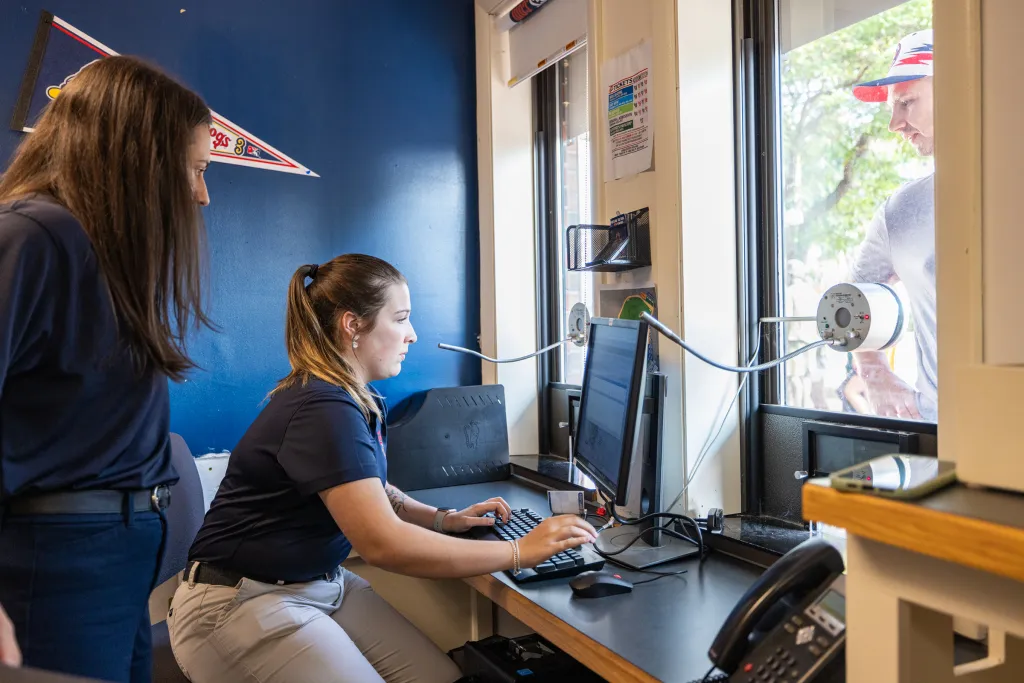 Two U N E students intern at a desk assisting a visitor through a window, with one seated at a computer and one standing nearby in a Sea Dog office.