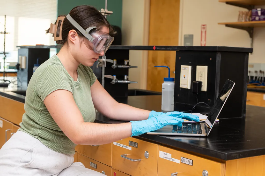 Science student wearing safety goggles and blue gloves working on a laptop at a laboratory bench with fume hood in the background.