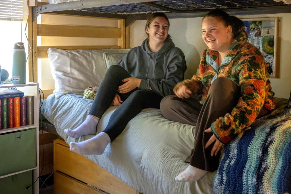 Two students sit on a lofted bed laughing together, one wearing a gray hoodie and the other in a colorful crocheted cardigan, with books visible on shelves below.