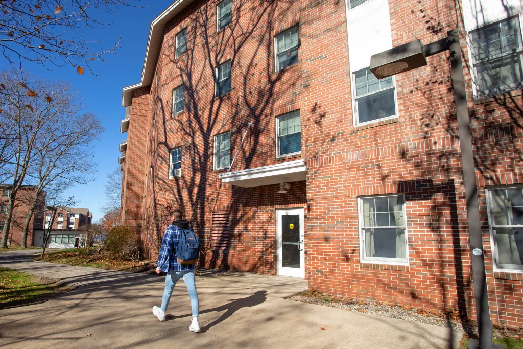 Student walks toward the entrance of a brick residence hall on a sunny autumn day with bare trees casting shadows on the building.