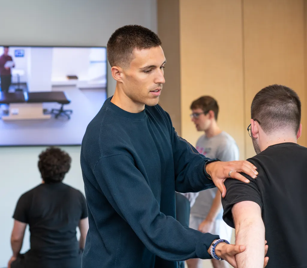 A UNE Doctor of Physical Therapy student performs a shoulder assessment on a classmate during a skills lab, with other student pairs practicing in the background.