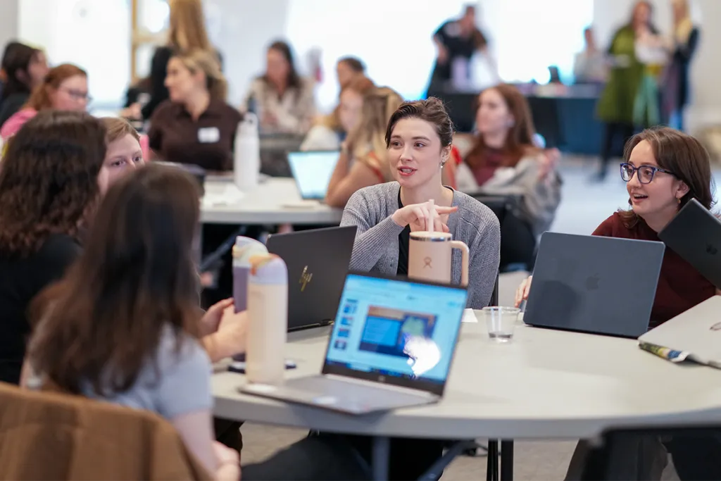 Students engage in small group discussion around a table with laptops open during a collaborative learning event.