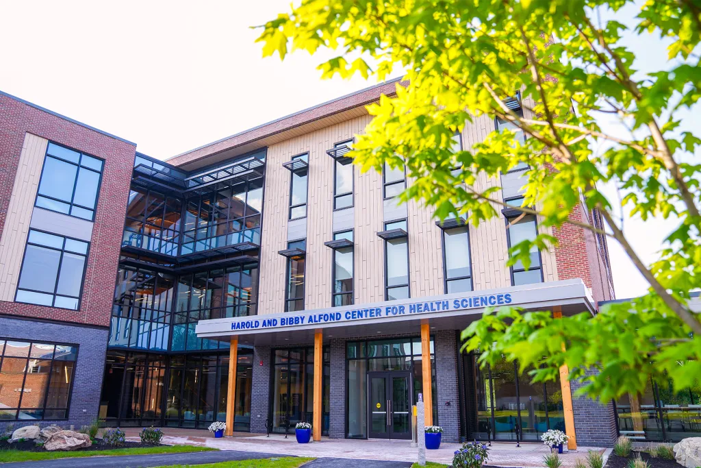 The exterior of the Harold and Bibby Alfond Center for Health Sciences on UNE's Biddeford campus, a modern multi-story building with floor-to-ceiling windows, wood accents, and blue flower planters at the entrance, framed by green summer foliage.