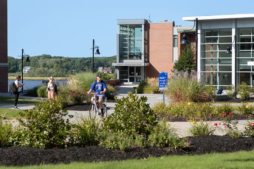Students walk and bike along a landscaped campus pathway on a sunny day, with the George and Barbara Bush Center visible in the background alongside a river view and flowering gardens.