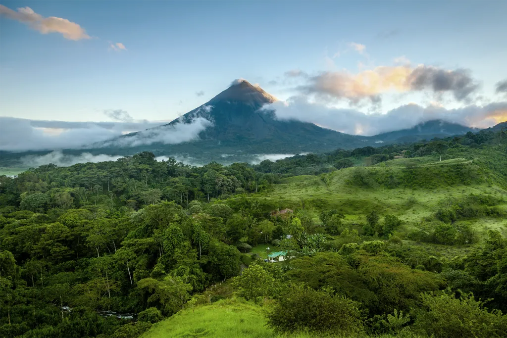 An aerial view of a lush Costa Rican rainforest with rolling green hills and a small dwelling nestled among the trees, set against the dramatic peak of Arenal Volcano rising through low clouds at dawn.