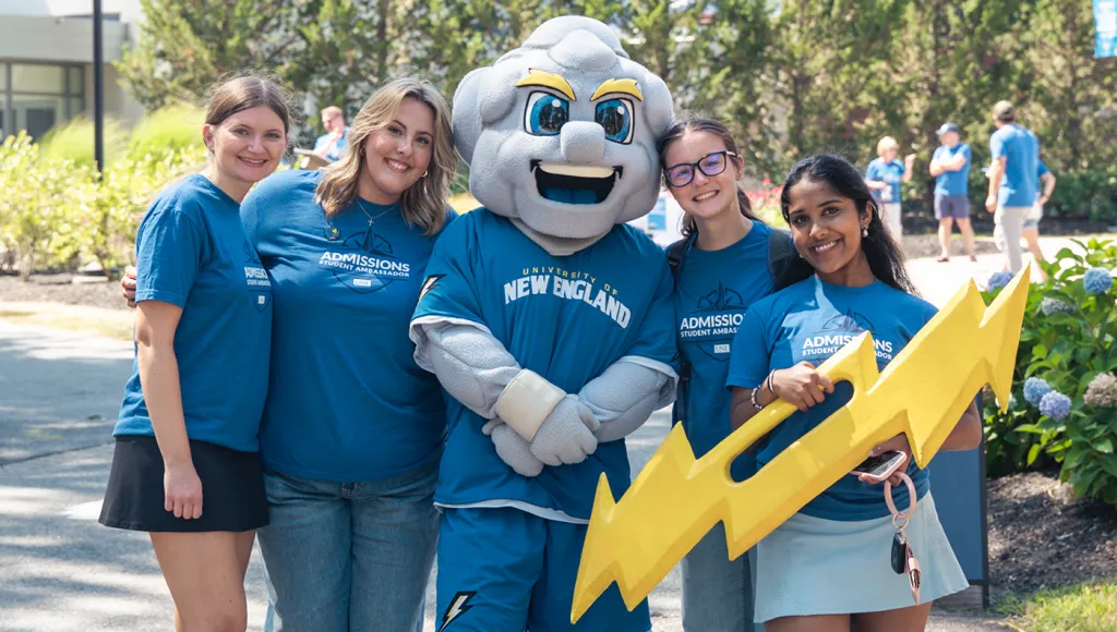 Four admissions student ambassadors pose with the U N E mascot holding a large yellow lightning bolt prop.