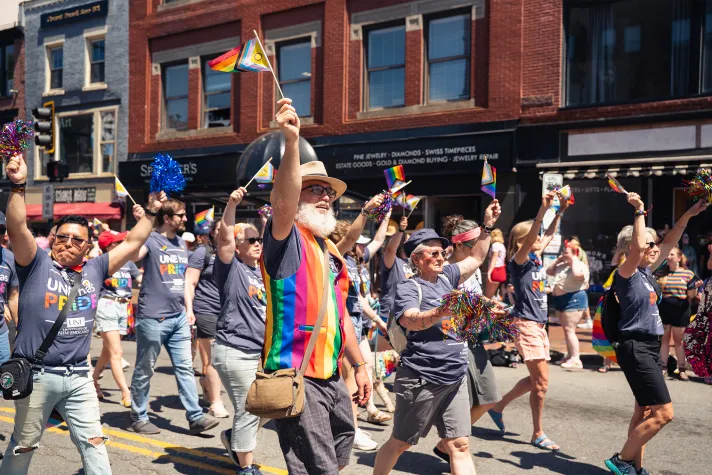 A group of employees in t-shirts that say, "U N E has Pride" walk together during a Pride Parade