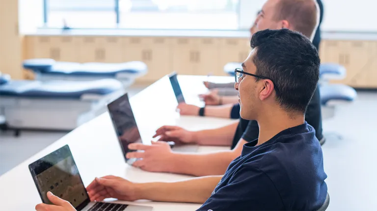 Two students in a classroom sit at a long table with their laptops open