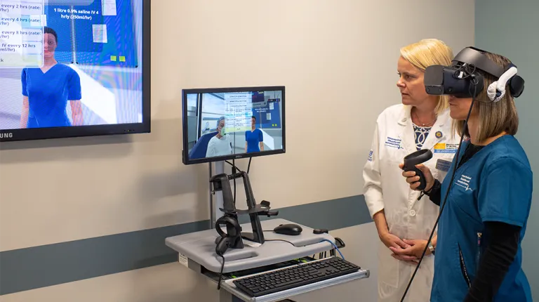 A student in a V R headset stands next to their professor as they review a Sim Lab experience on a monitor