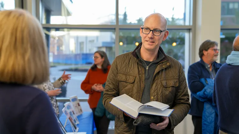 Eric Zuellow looking at a book and chatting with someone in the foreground of the image