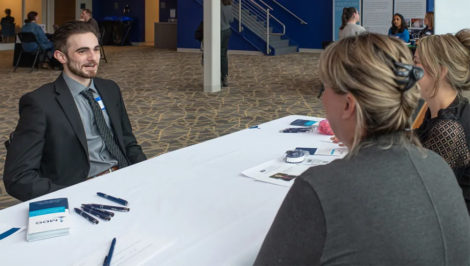 A student wearing a suit and tie performs a mock interview with two interviewers