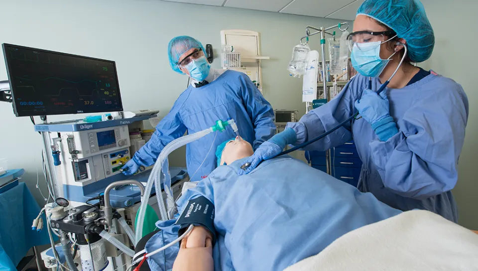 An osteopathic medicine student and professor in scrubs work on a patient simulator in U N E's sim lab surgery room