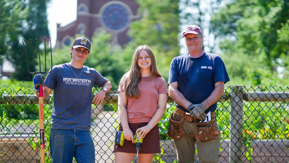 Luke Jenkins (Biology, ’26), Cameron Wake, Ph.D., and a student pose with gardening tools and power tools in front of a chain link fence.