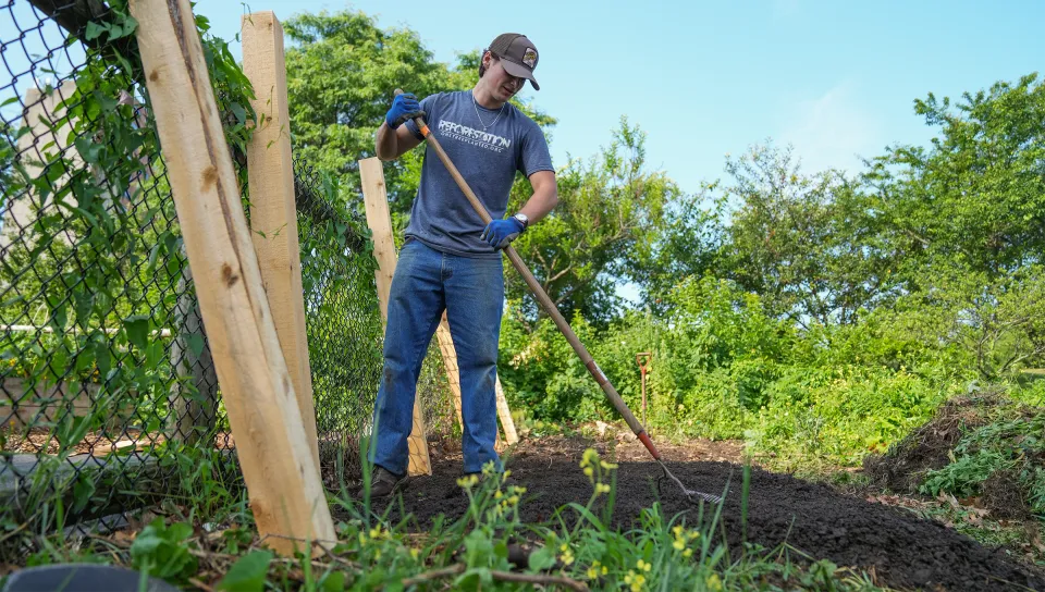 Luke Jenkins (Biology, ’26) digs into a dirt mound in a community garden with greenery in the background