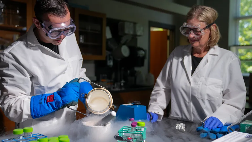 A graduate biology student and their professor in lab coats and safety goggles handle a smoking container of liquid nitrogen while conducting cryogenic laboratory work with sample vials and testing equipment.