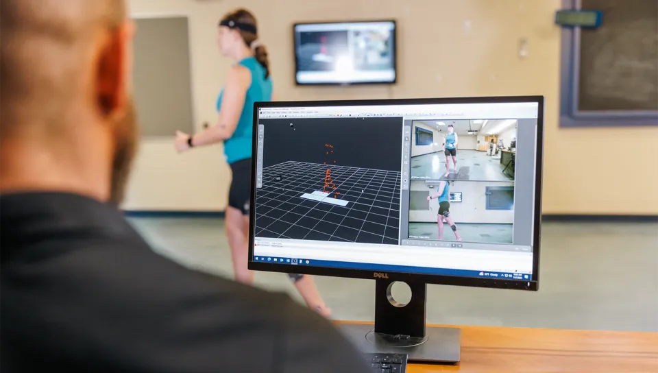 A professor monitors motion capture data on a computer screen as a student participant wearing sensor markers walks through a biomechanics lab in the background. The screen displays a 3D skeletal model alongside multi-angle video feeds of the participant.