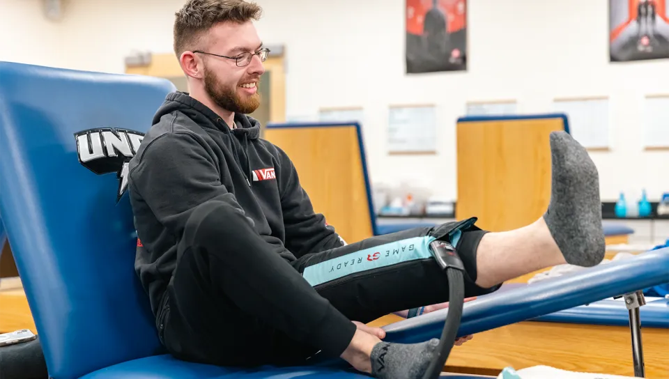 A UNE student reclines on a treatment table in the athletic training room with a Game Ready compression and cold therapy wrap applied to his lower leg.