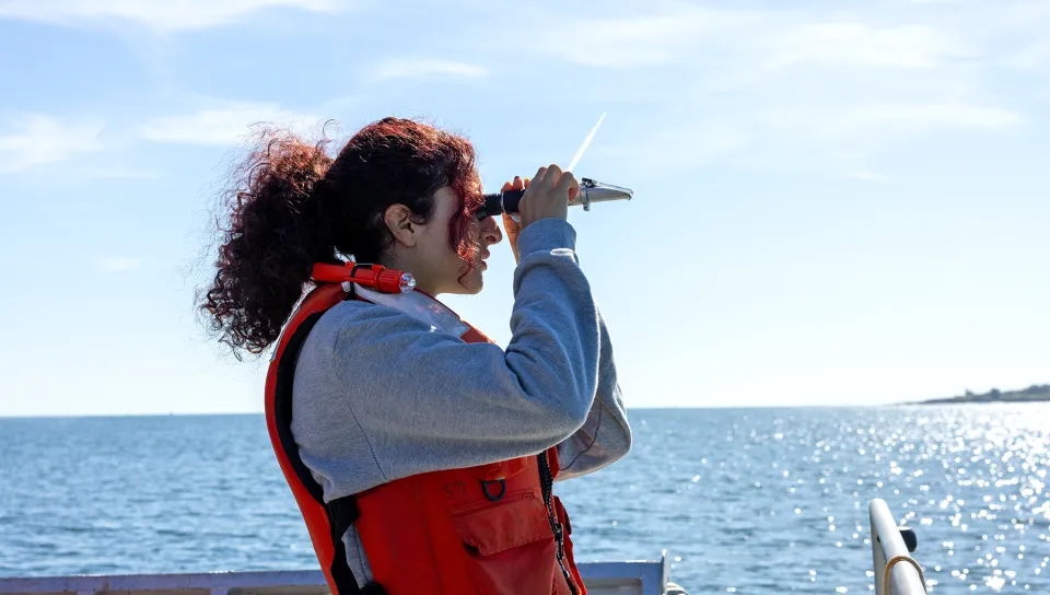 A UNE marine sciences student wearing an orange life jacket looks through a handheld scope from the deck of a research vessel on open water.