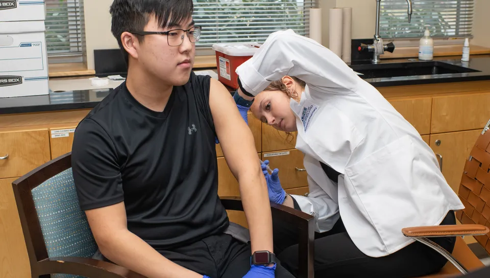 A UNE pharmacy student in a white coat and gloves administers an intramuscular injection into the upper arm of a classmate during a vaccine administration skills lab.