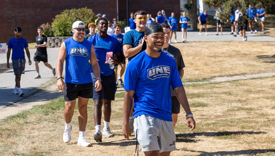 A large group of UNE Nor'easters students in blue UNE shirts walk together across campus on a sunny day, smiling and laughing during move-in festivities.