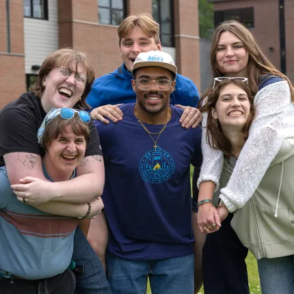 Group of six students pose together outdoors on campus, smiling and embracing in casual spring clothing.