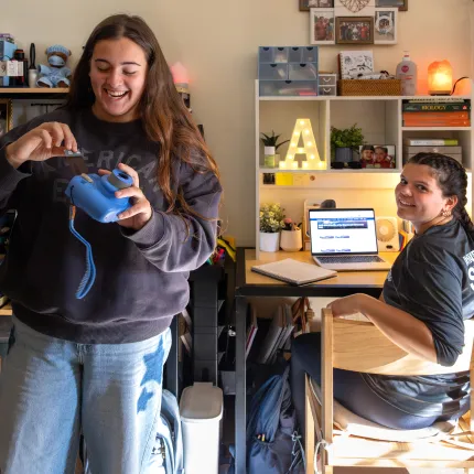 Two students laugh together in a cozy, personalized dorm room. One stands holding a blue toy camera while the other sits at a desk with an open laptop, surrounded by shelves decorated with photos, plants, a glowing letter light, and a Himalayan salt lamp.