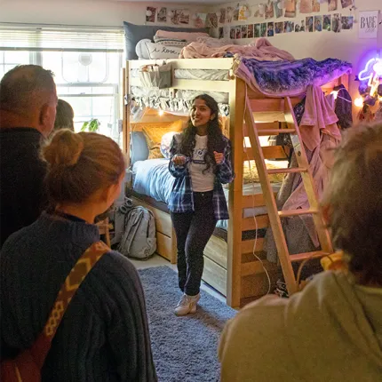 A student shows visitors her decorated dorm room during a campus tour, standing beside a lofted bed adorned with photos, string lights, and a neon sign.