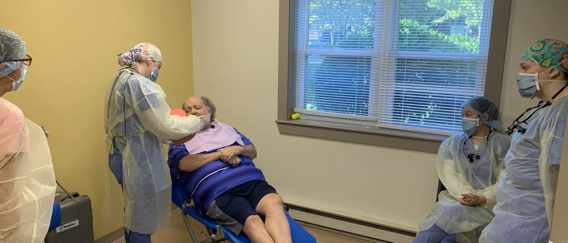 A patient, seated in a dentist chair, is consulted by students